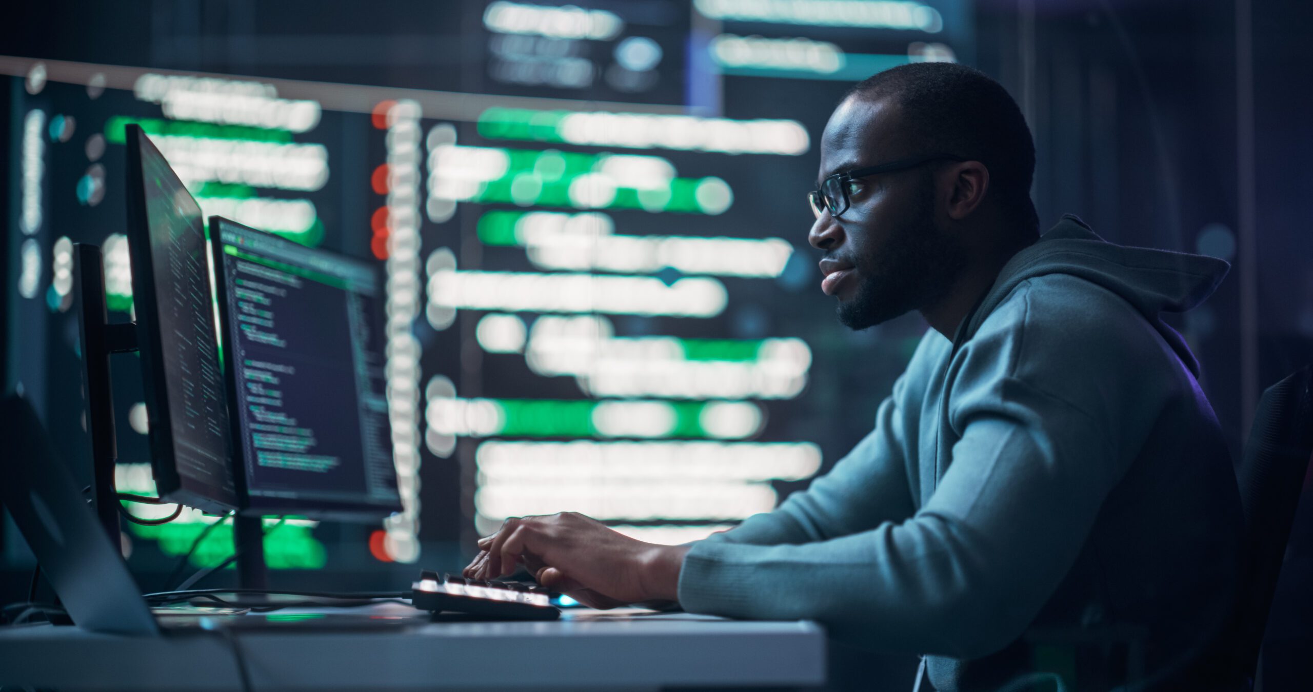 Focused Black Male Programmer Working in Monitoring Room, Surrounded by Big Screens Displaying Lines of Programming Language Code. Portrait of Man Creating Software. Futuristic Coding Concept.
